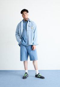 Lyseblå denimjakke over en matchende skjorte, parret med knælange blå shorts. Sorte sneakers med grønne accenter. Hvid baggrund.