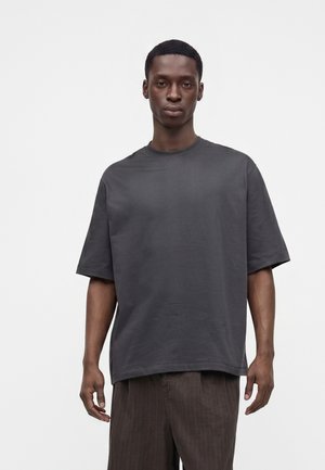 Homme aux cheveux courts portant un t-shirt ample gris foncé à manches courtes et un pantalon marron, debout devant un fond clair uni.