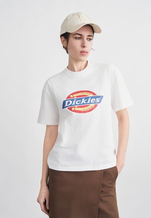 Naine, kes kannab beežit LA-mütsi, valget Dickies'i logoga t-särki ja pruuni seelikut, seisab valge tausta ees.