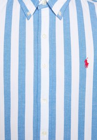 Modrobíle pruhovaná košile na knoflíky vyrobená z bavlny, s límcem, knoflíkovou přední částí a malým červeným logem na levé straně hrudi.