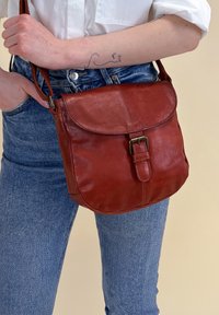 Hnědá kožená crossbody taška se sklápěcím víkem a kovovou přezkou. Hladký povrch s zaoblenými okraji, nošená přes džíny.