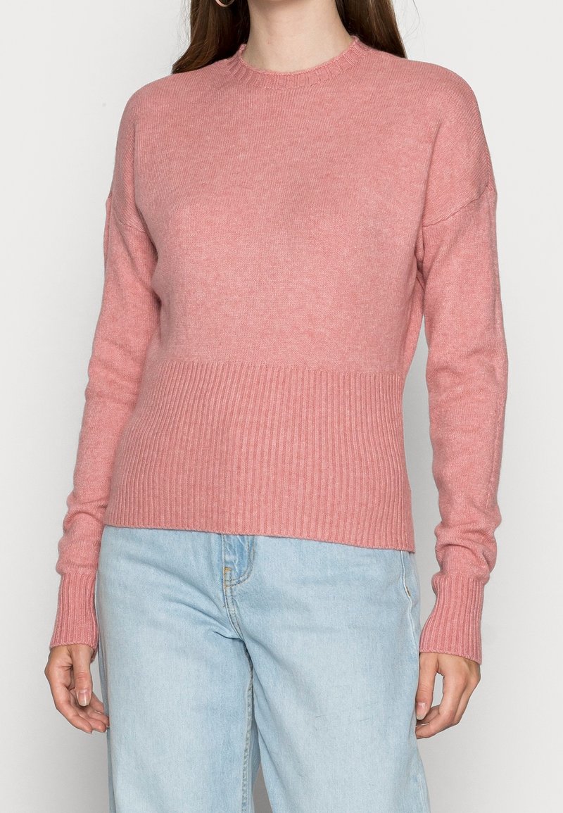 Kvinna iklädd en långärmad rosa stickad tröja med ribbad nederkant och ärmslut, tillsammans med ljusblå denimjeans, mot en enhetlig ljus bakgrund.