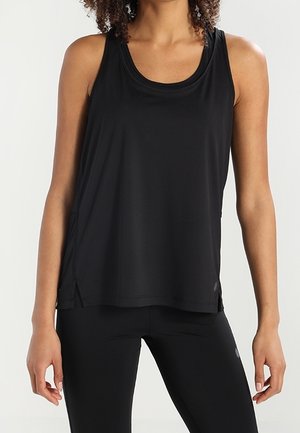 Frau, die ein ärmelloses schwarzes Sporttop und schwarze Leggings trägt, steht mit entspannten Armen vor einem einfarbigen Hintergrund.