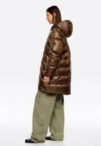 Brązowa pikowana kurtka puchowa z kapturem, błyszczącą fakturą i poziomym pikowaniem. Połączona z luźnymi zielonymi spodniami i czarnym obuwiem.