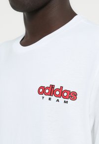 Λευκό βαμβακερό t-shirt με στρογγυλή λαιμόκοψη, διακοσμημένο με κόκκινο και μαύρο λογότυπο "adidas TEAM" στο αριστερό μέρος του στήθους. Ομαλή υφή.