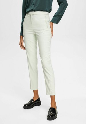 Vrouw die roomkleurige, enkellange, op maat gemaakte broek draagt, donkergroene blouse met lange mouwen en glanzende zwarte loafers, staand tegen een witte achtergrond.