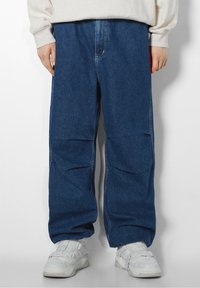 Baggy blå denimjeans med en slät yta, med en midjehöjd som är måttlig och stora sidofickor, tillsammans med vita sneakers.
