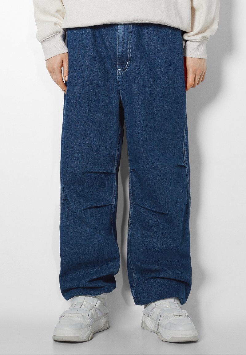 Baggy blå denimjeans med en slät yta, med en midjehöjd som är måttlig och stora sidofickor, tillsammans med vita sneakers.