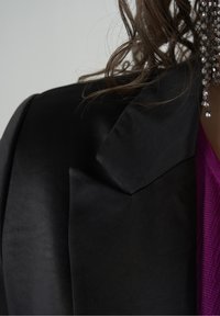 Blazer de satén negro con cuello con muesca, combinado con una prenda texturizada púrpura por debajo. Joyería brillante visible.