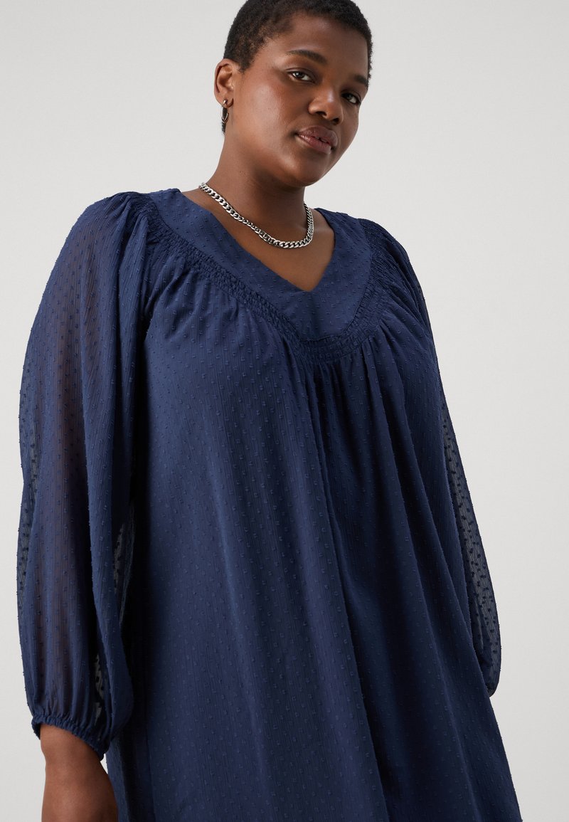 Robe texturée bleu foncé avec un décolleté en V, des détails froncés et des manches volumineuses. Fabriquée dans un tissu doux et léger avec un motif subtil.