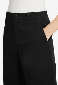 Pantalons noirs avec une fermeture à bouton, une poche latérale et un tissu lisse et texturé. La taille est élastique pour plus de confort.