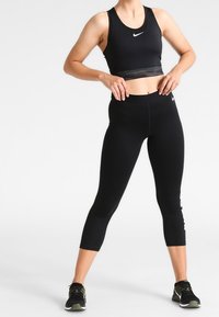 Zwarte sportcrop top met een naadloos ontwerp, gecombineerd met hoge zwarte leggings. Bevat het witte Nike-logo en accent op het been.
