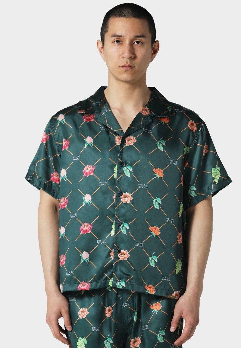 Camisa verde de manga corta hecha de tejido satinado, con patrones florales en rosa, naranja y verde, con un cierre de botones en la parte delantera y un corte relajado.