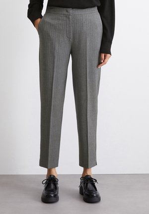 Pantaloni gri, croiți pe măsură, cu un model cu dungi fine, tăiere dreaptă și mici slituri laterale la tiv, asortați cu pantofi negri cu șireturi.