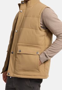 Chaleco acolchado beige con botones a presión y un gran bolsillo frontal, llevado sobre un suéter de manga larga marrón claro y pantalones oscuros.