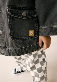 Veste en denim noire avec des accessoires en argent, arborant une étiquette de visage souriant. Associée à un pantalon à carreaux noir et blanc et des baskets noires.