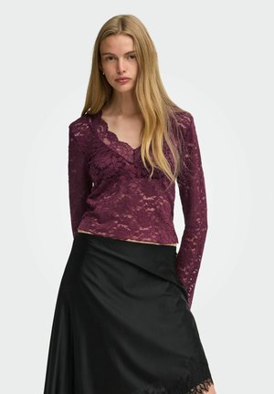 Femme aux longs cheveux blonds portant un haut en dentelle bordeaux à manches longues et une jupe en satin noire sur un fond clair uni.