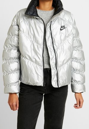 Persoon draagt een glanzend zilveren Nike pufferjack over een grijze trui en zwarte jeans, staand met ontspannen armen langs het lichaam.