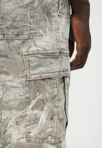 Világosszürke anyagú cargo nadrág, fanyaga mintázattal, oldalsó zsebbel és látható varrási részletekkel. Sima textúra.