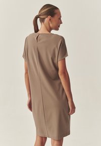 Krátké šaty s krátkým rukávem délky ke kolenům v měkké taupe látce. Mají zadní výstřih na knoflík a rovný střih. Hladká textura.