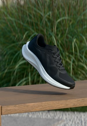 Schwarzer Sportschuh mit weißer Sohle schwebt über einer Holzbank, im Hintergrund grünes hohes Gras.