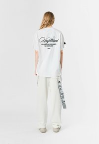 Camiseta blanca de manga corta con texto negro en la parte de atrás que dice "Sin ataduras" y una frase debajo. Combinada con pantalones blancos sueltos.