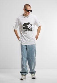 White oversized cotton t-shirt with a black distressed "STAR" logo, paired with light blue baggy jeans and white sneakers with accents.