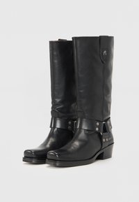 Botas de cuero negro hasta la rodilla con punta curva, correa y tachuelas metálicas alrededor del tobillo, y un tacón pequeño. Presenta un logo cerca de la parte superior.