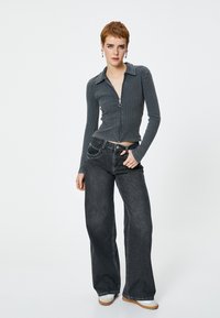 Mørkegrå ribbet zip-up trøje med krave, parret med vide mørke denimbukser. Modellen har hvide sneakers med en tan sål på.