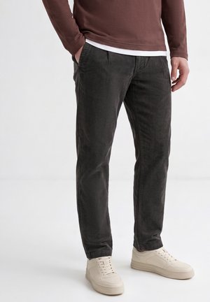 Homme portant un pantalon en velours côtelé gris foncé, une chemise marron à manches longues et des baskets blanches, debout sur un fond blanc uni.