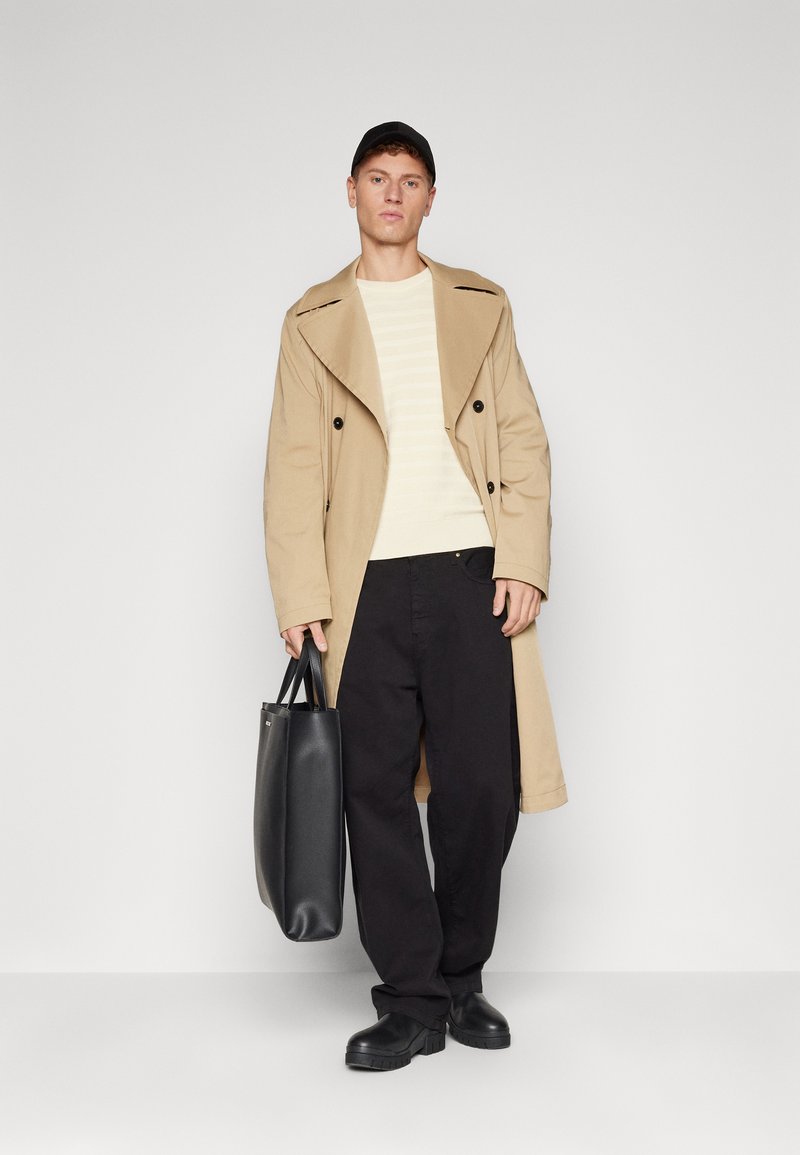 Beżowy płaszcz trench z podwójnym zapięciem, noszony na lekki sweter w paski, w zestawie z czarnymi spodniami i czarną torbą na ramię.