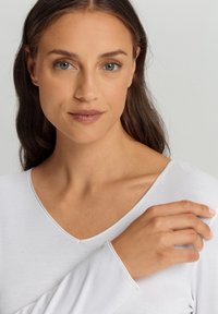 Weißes Langarmoberteil mit V-Ausschnitt. Der Stoff wirkt weich und hat eine glatte Textur. Die Hand des Models ruht auf der Schulter und zeigt den natürlichen Nagel.