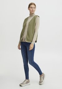 Olivno zeleni flis brezrokavnik z zadrgo spredaj, nanešen čez bež polo majico. Nositi z modrimi ožjimi jeans hlačami in sivimi supergami z detajli.