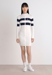 Abito bianco a coste con righe orizzontali blu navy, maniche lunghe e una cerniera lampo frontale completa. Abbinato a sneakers bianche e calzini.