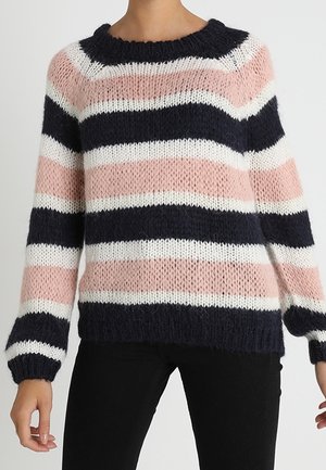 Femme portant un pull en maille ample à rayures horizontales bleu marine, rose et blanc, associé à un pantalon noir.