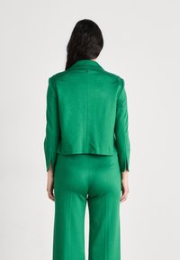 Short green jacket with a structured collar, tailored fit, and back slit detail. Paired with matching wide-leg trousers.