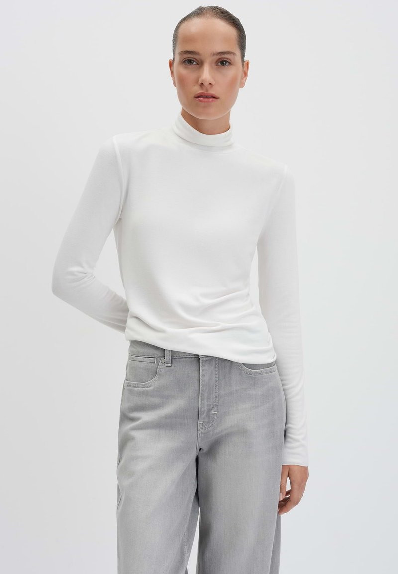 Langärmliges weißes Rollkragenoberteil aus weichem Stoff, kombiniert mit hellgrauen Jeans; hat einen lässigen Schnitt und eine glatte Textur.