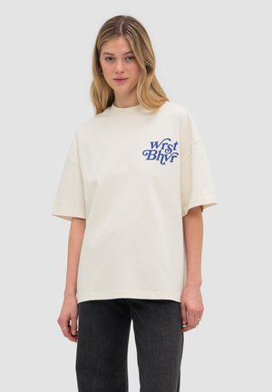 Młoda kobieta z długimi blond włosami, ubrana w kremową, oversize'ową koszulkę z niebieskim napisem na piersi i ciemne spodnie, stoi.