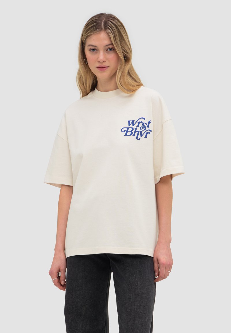 Junge Frau mit langen blonden Haaren, die ein cremeweißes übergroßes T-Shirt mit blauem Text auf der Brust und dunkle Hosen trägt, stehend.