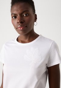 White cotton t-shirt with short sleeves, featuring a subtle tonal embroidered flower design on the left side and a round neckline.