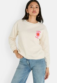 Femme portant un sweat crème avec un graphique de soleil rose et le texte "Good Vibes", associée à un jean bleu clair, posant sur un fond blanc.