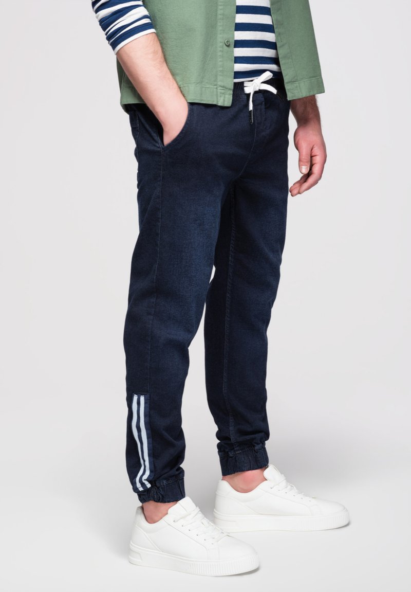 Mann in marineblauen Jogginghosen mit Seitenstreifen, weißen Sneakers, gestreiftem Hemd und grüner Jacke, der mit einer Hand in der Tasche steht.