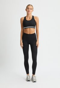 Reggiseno sportivo atletico nero con una fascia con logo, abbinato a leggings neri con un motivo geometrico testurizzato. Indossato con scarpe da ginnastica argentate.