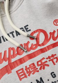 Grijze hoodie met rode en zwarte tekst. Crème kleurige koordjes met metalen uiteinden. Stof toont een lichte textuur in het afdrukontwerp.