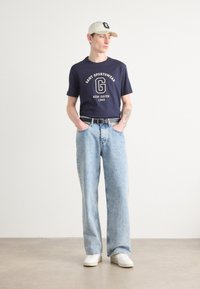 Marine T-skjorte med hvitt trykk, lyseblå baggy jeans og hvite joggesko. Lys khaki caps med logo. Uformell stil med synlig tatovering på armen.
