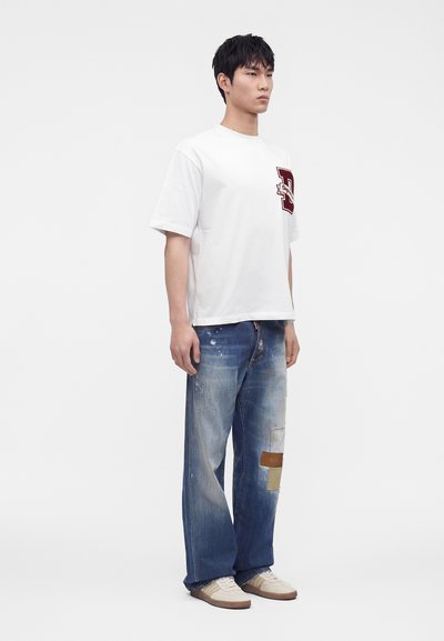 Νεαρός άνδρας που φοράει λευκό υπερμεγέθη T-shirt με κόκκινο πάτκι, φαρδιά μπλε τζιν με πάτκες και μπεζ αθλητικά παπούτσια, στέκεται μπροστά σε απλό φόντο.