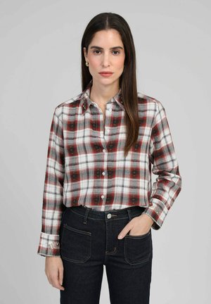 Femme aux cheveux longs et foncés portant une chemise à carreaux rouge et blanche boutonnée, rentrée dans un jean foncé, debout devant un fond gris.