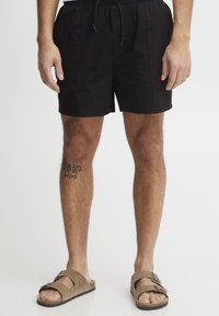 Mustad ujumispüksid elastse vöökohaga ja paelaga, sirge lõikega. Kandmisel pruunide sandaalidega, nähtav jala tätoveering.