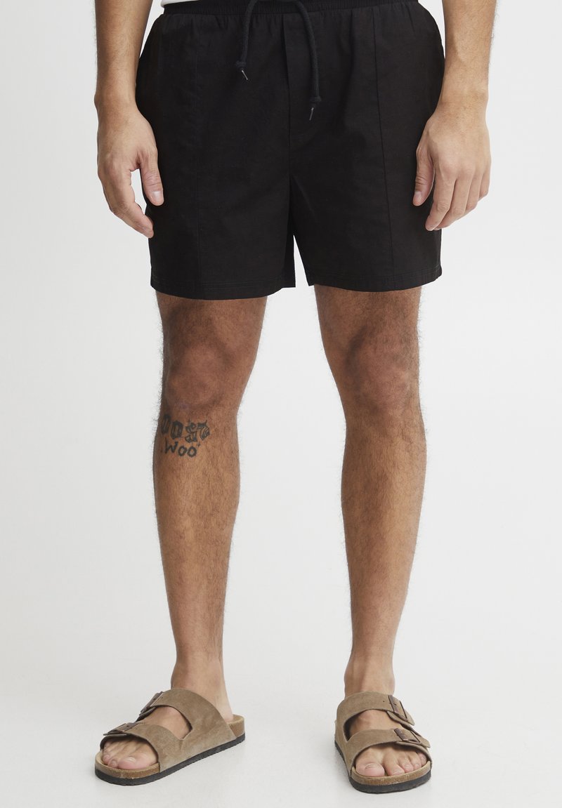 Black swim shorts with an elastic waistband and drawstring, featuring a straight cut. Worn with brown sandals, showing a leg tattoo.