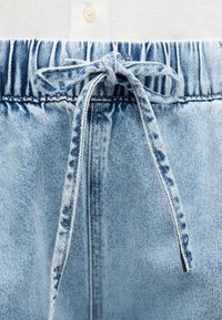 Φωτεινό γαλάζιο τζιν παντελόνι με ελαστική μέση και δεμένο κορδόνι τζιν, πάνω σε λευκό ριγωτό ύφασμα.